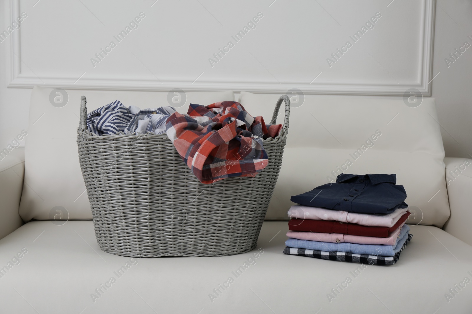 Photo of Wicker basket with dirty laundry and stack of clean clothes on sofa indoors