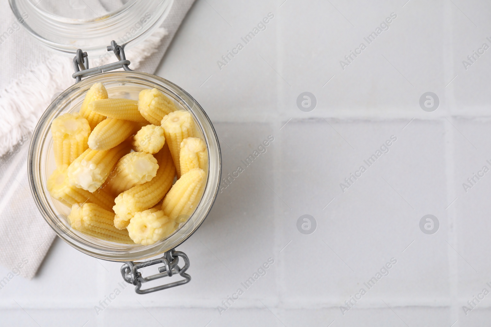 Photo of Tasty fresh yellow baby corns in glass jar on white tiled table, top view. Space for text