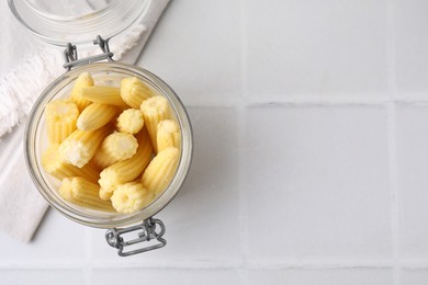 Tasty fresh yellow baby corns in glass jar on white tiled table, top view. Space for text