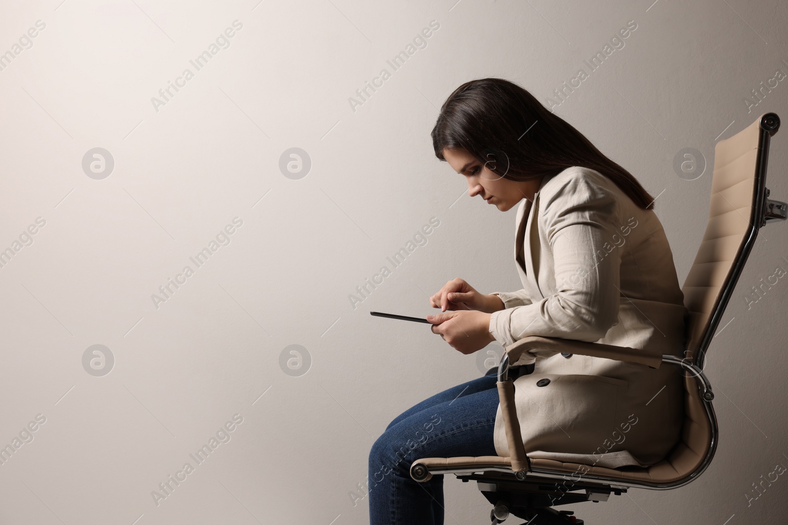 Photo of Young woman with bad posture using tablet while sitting on chair against grey background. Space for text