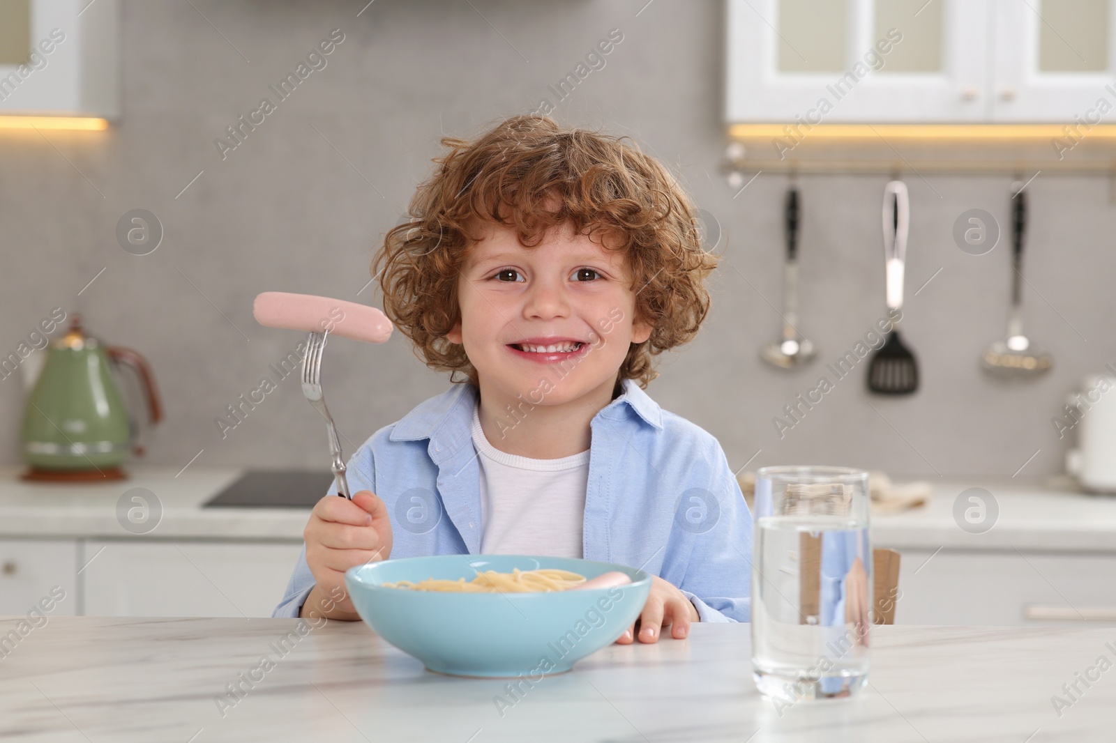 Photo of Cute little boy holding fork with sausage and bowl of pasta at table in kitchen