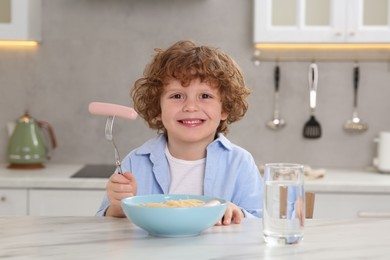 Photo of Cute little boy holding fork with sausage and bowl of pasta at table in kitchen