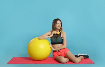 Beautiful woman with headphones sitting on yoga mat near fitness ball against turquoise background