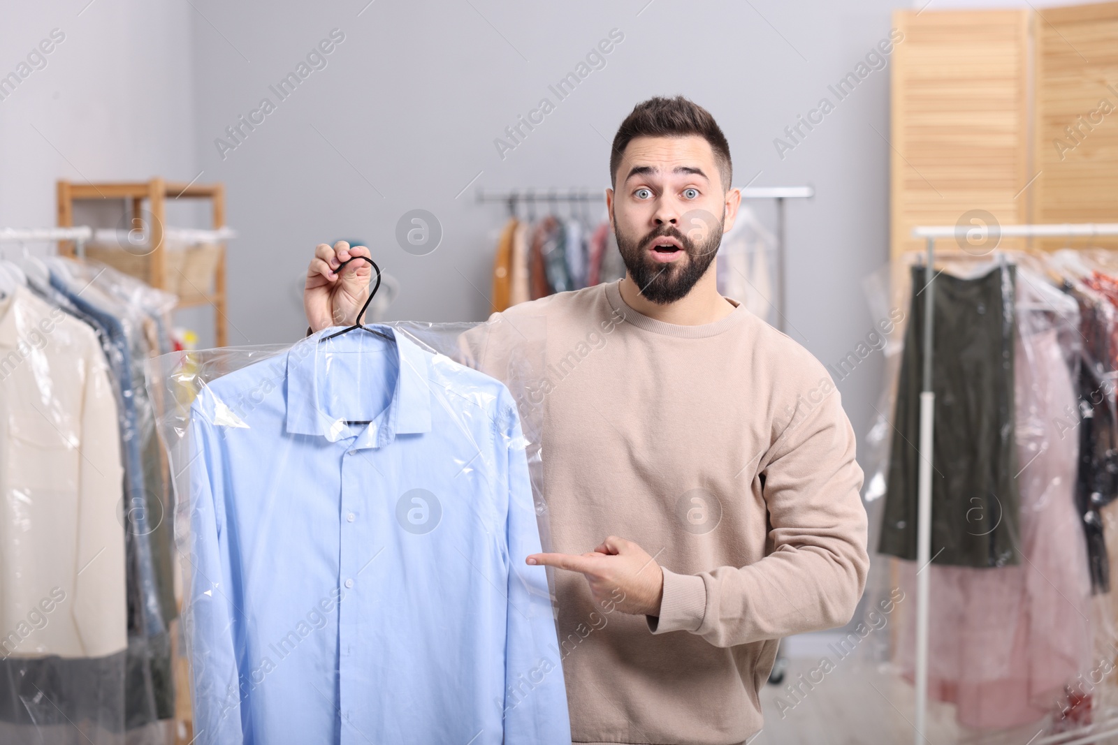 Photo of Dry-cleaning service. Shocked man holding hanger with shirt in plastic bag indoors