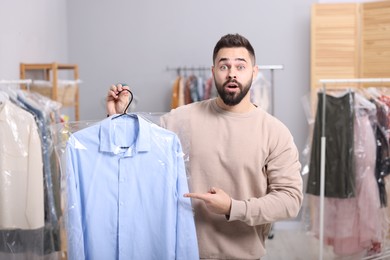 Dry-cleaning service. Shocked man holding hanger with shirt in plastic bag indoors