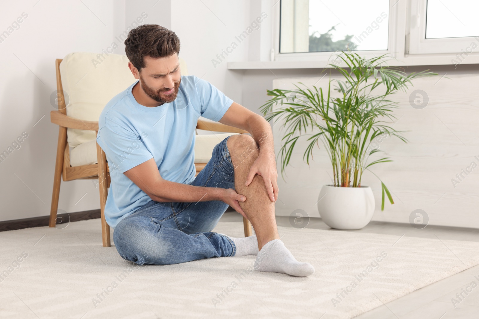 Photo of Man suffering from leg pain on white carpet near armchair at home