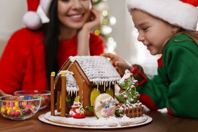 Photo of Mother and daughter with gingerbread house at table indoors, closeup