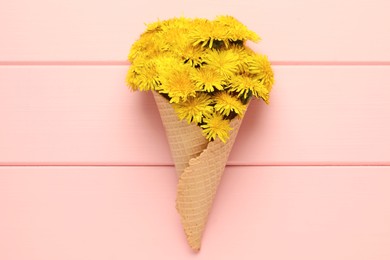 Photo of Beautiful yellow dandelions in waffle cone on pink wooden table, top view
