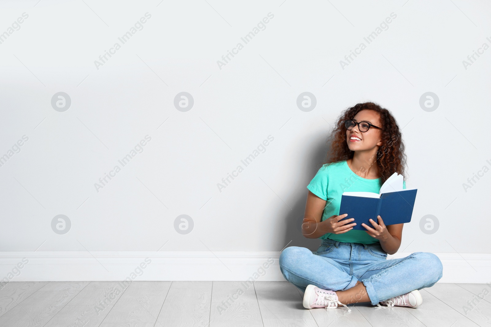 Photo of Beautiful young African-American woman reading book on wooden floor near light wall, space for text
