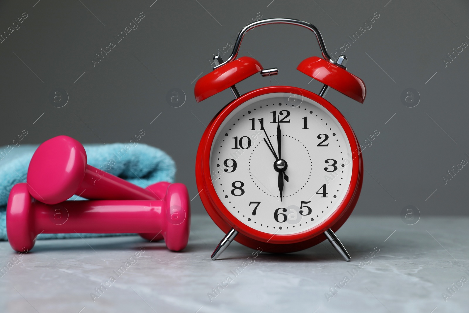 Photo of Alarm clock, dumbbells and towel on table against grey background. Morning exercise