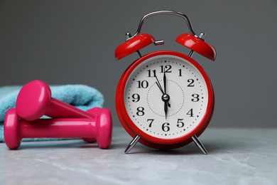 Photo of Alarm clock, dumbbells and towel on table against grey background. Morning exercise