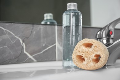 Photo of Natural loofah sponge and bottle with shower gel on washbasin in bathroom, closeup. Space for text