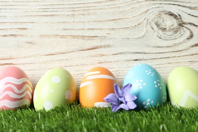 Colorful painted Easter eggs with flowers on green grass against wooden background, space for text