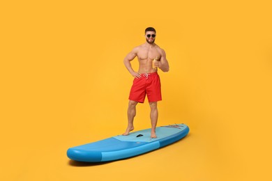 Photo of Man with refreshing drink posing on SUP board against orange background