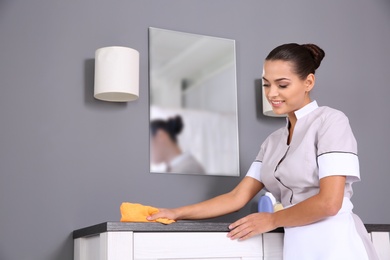 Photo of Young chambermaid wiping dust from furniture with rag in bathroom