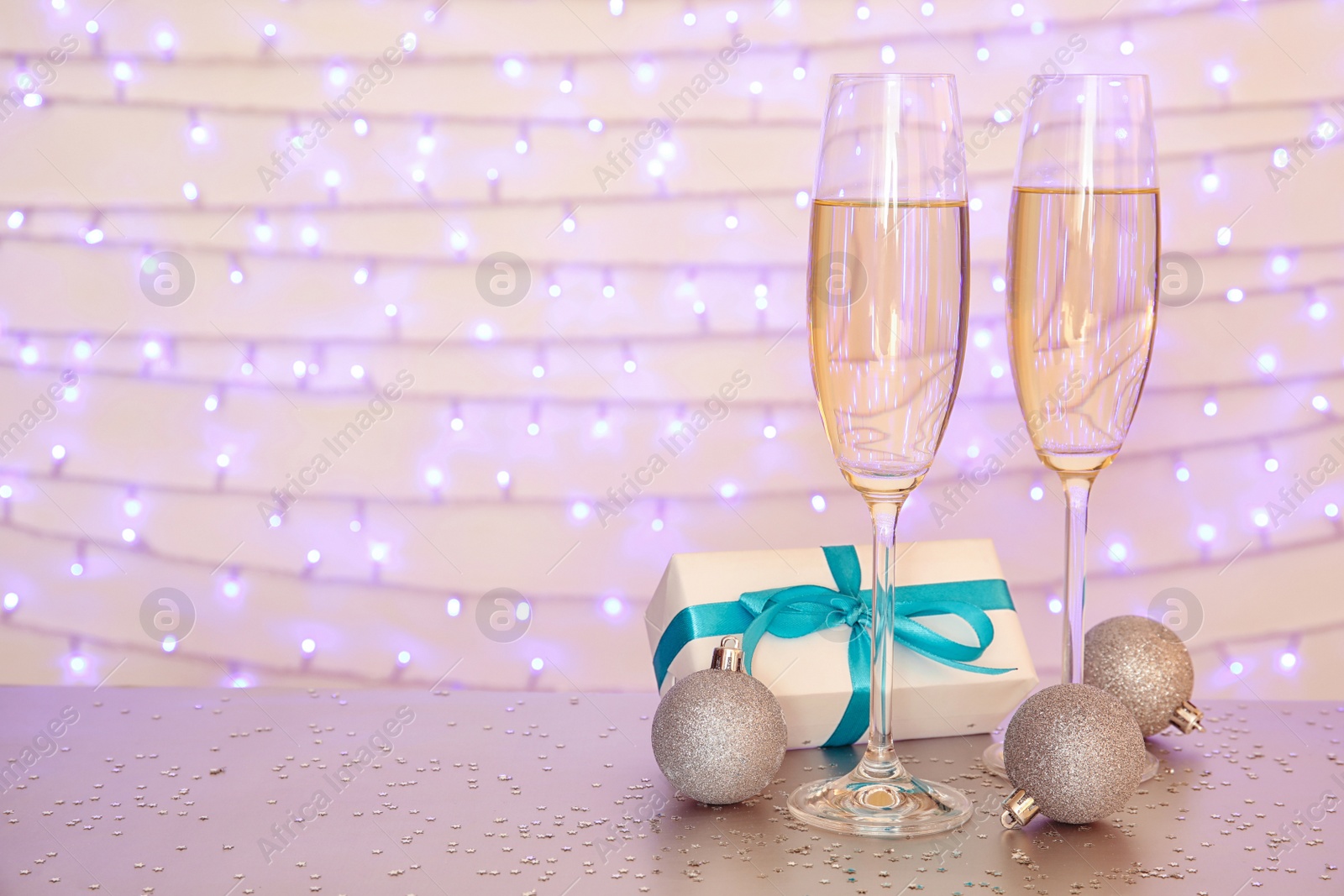 Photo of Glasses of champagne, decorative balls and Christmas gift on table against blurred lights