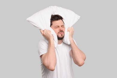 Photo of Tired man covering ears with pillow on light grey background. Insomnia problem