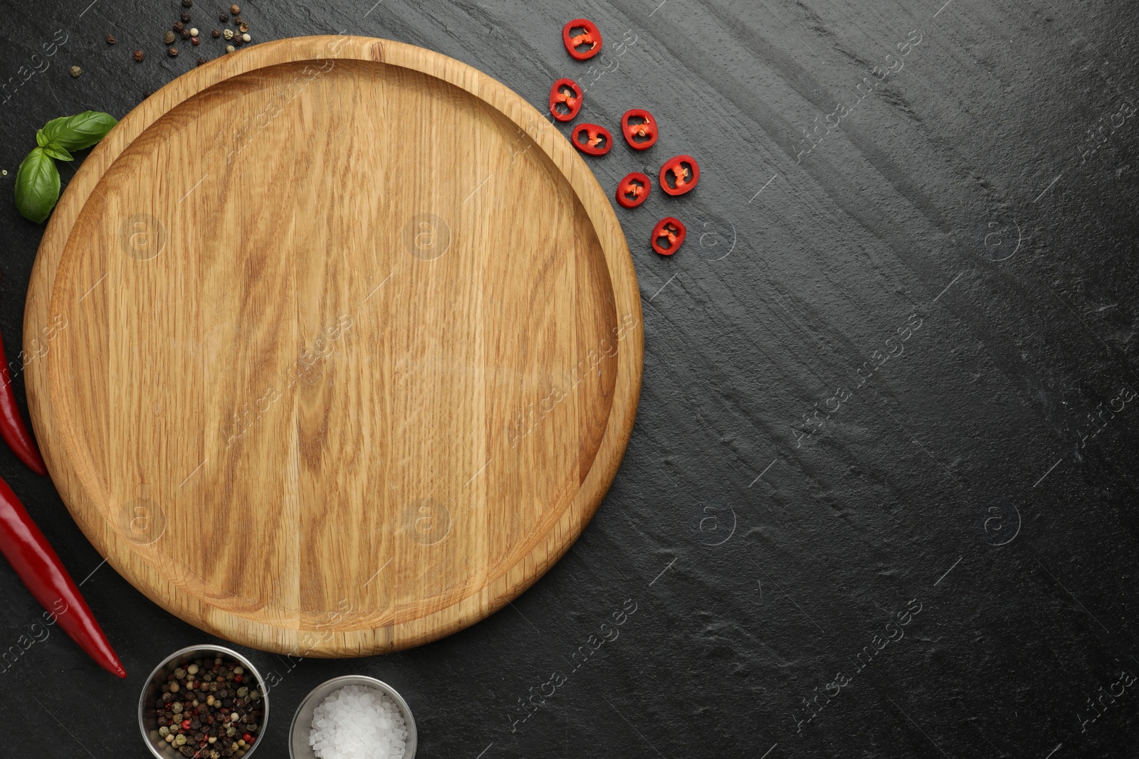 Photo of Cutting board, salt, spices, basil and chili peppers on black textured table, flat lay. Space for text