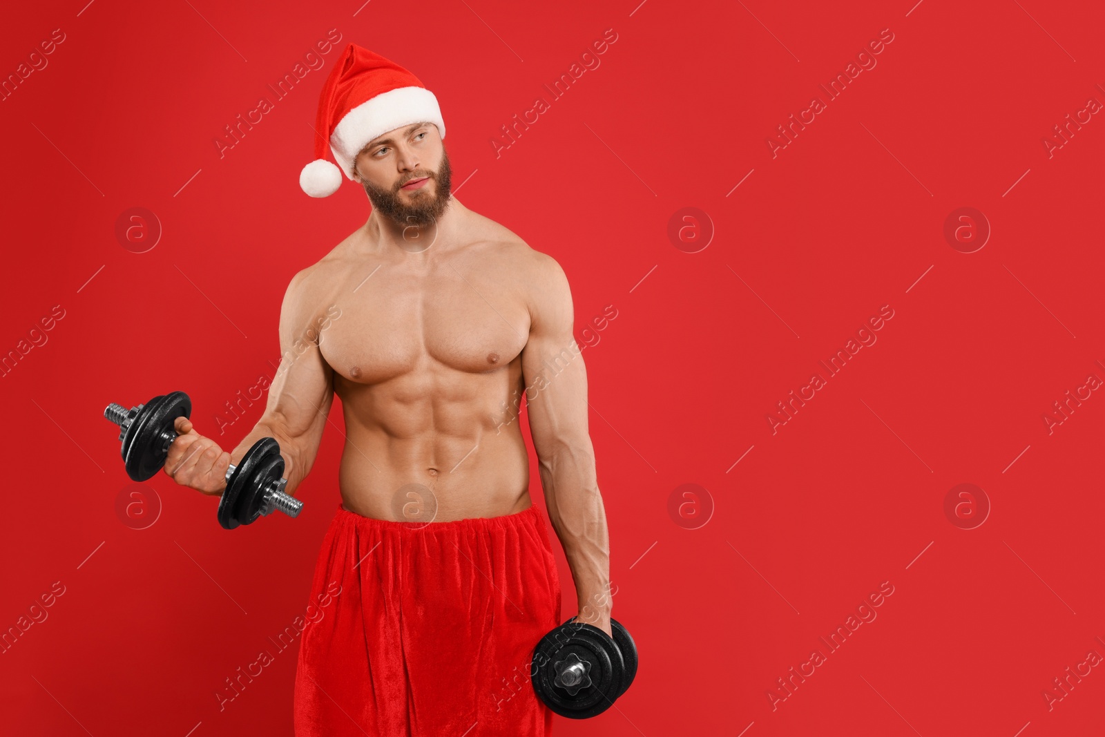 Photo of Muscular young man in Santa hat with dumbbells on red background, space for text