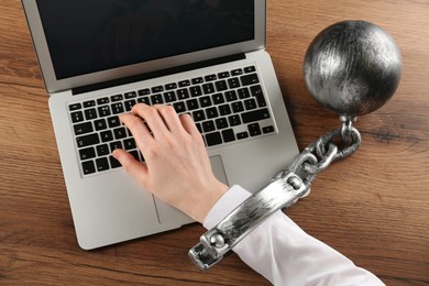 Photo of Woman shackled with ball and chain typing on laptop at wooden table, above view. Internet addiction