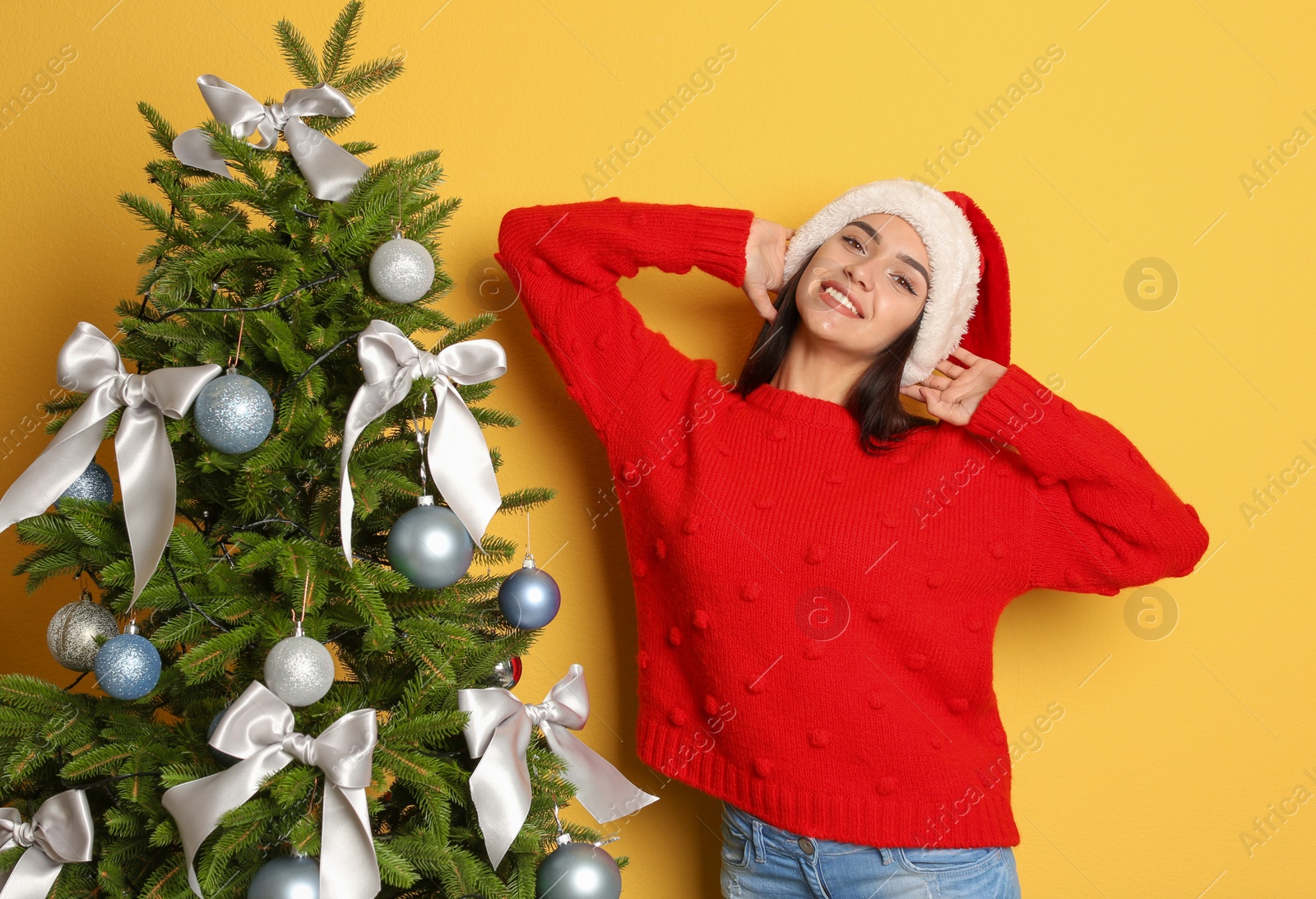 Photo of Beautiful young woman in Santa hat near Christmas tree on color background