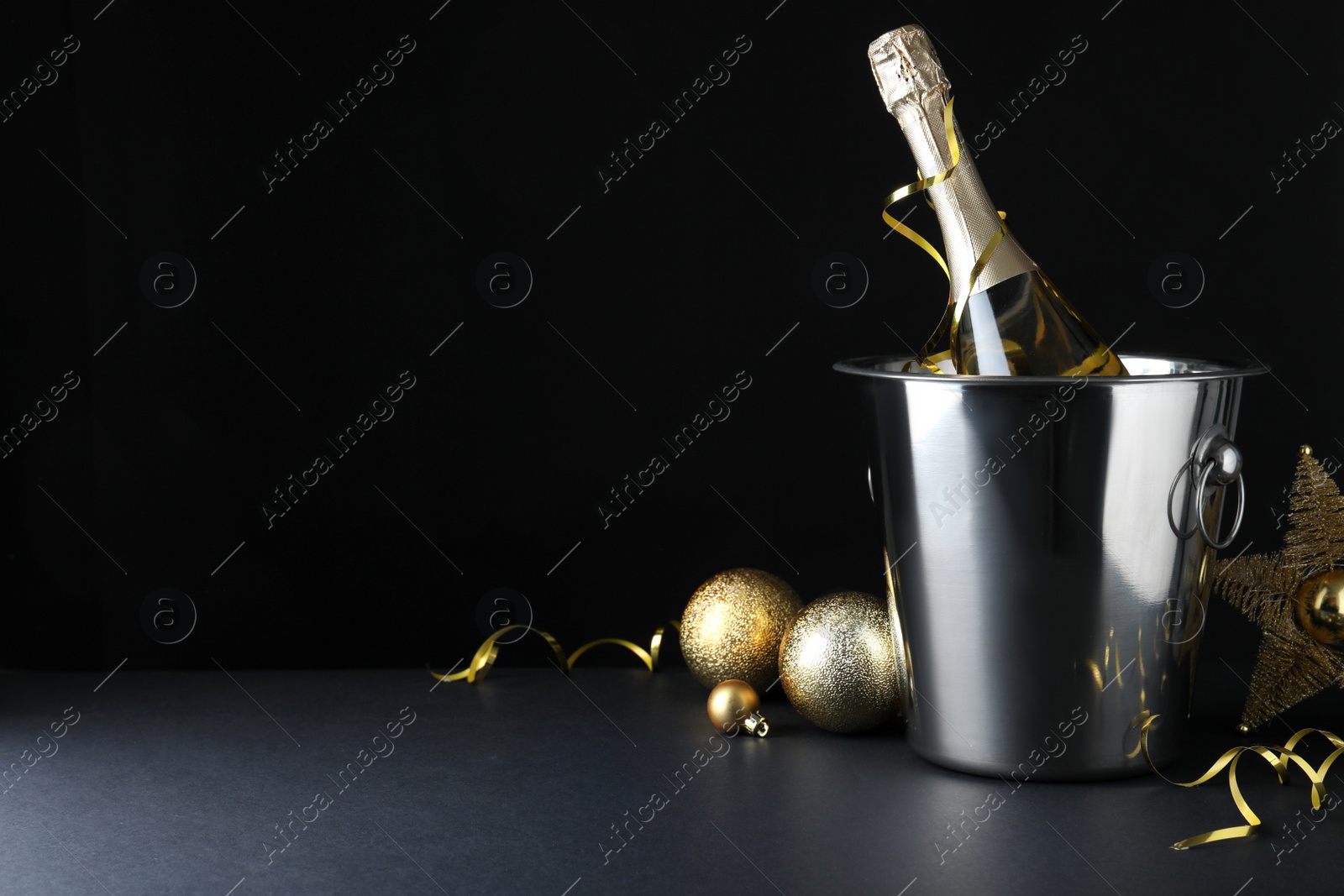 Photo of Happy New Year! Bottle of sparkling wine in bucket and festive decor on table against black background, space for text