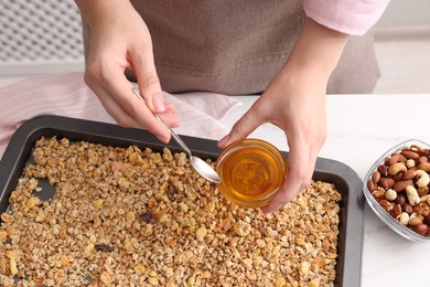 Photo of Making granola. Woman adding honey onto baking tray with mixture of oat flakes and other ingredients at white table in kitchen, closeup