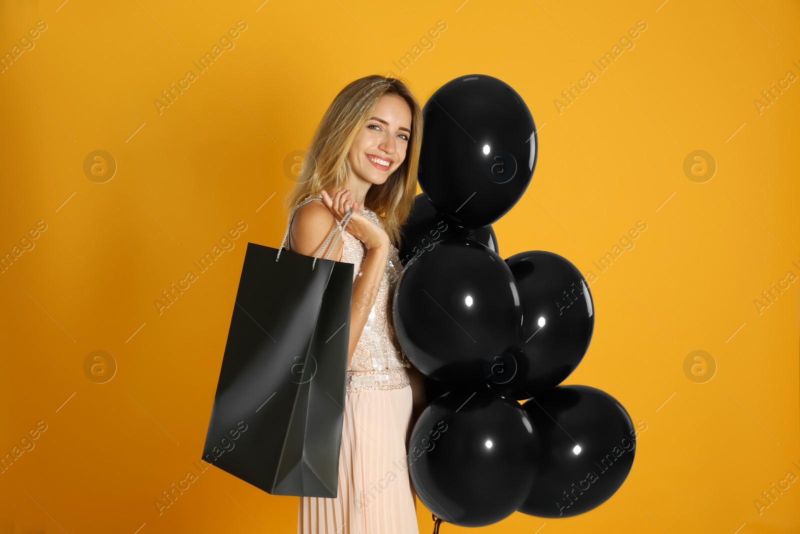 Photo of Happy young woman with balloons and shopping bag on yellow background. Black Friday Sale