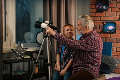 Photo of Little boy with his grandfather using telescope to look at stars in room