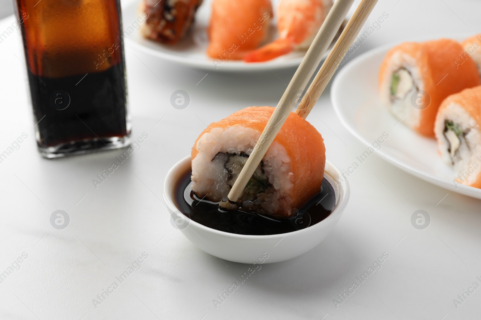 Photo of Dipping tasty sushi roll with salmon into soy sauce on white table, closeup