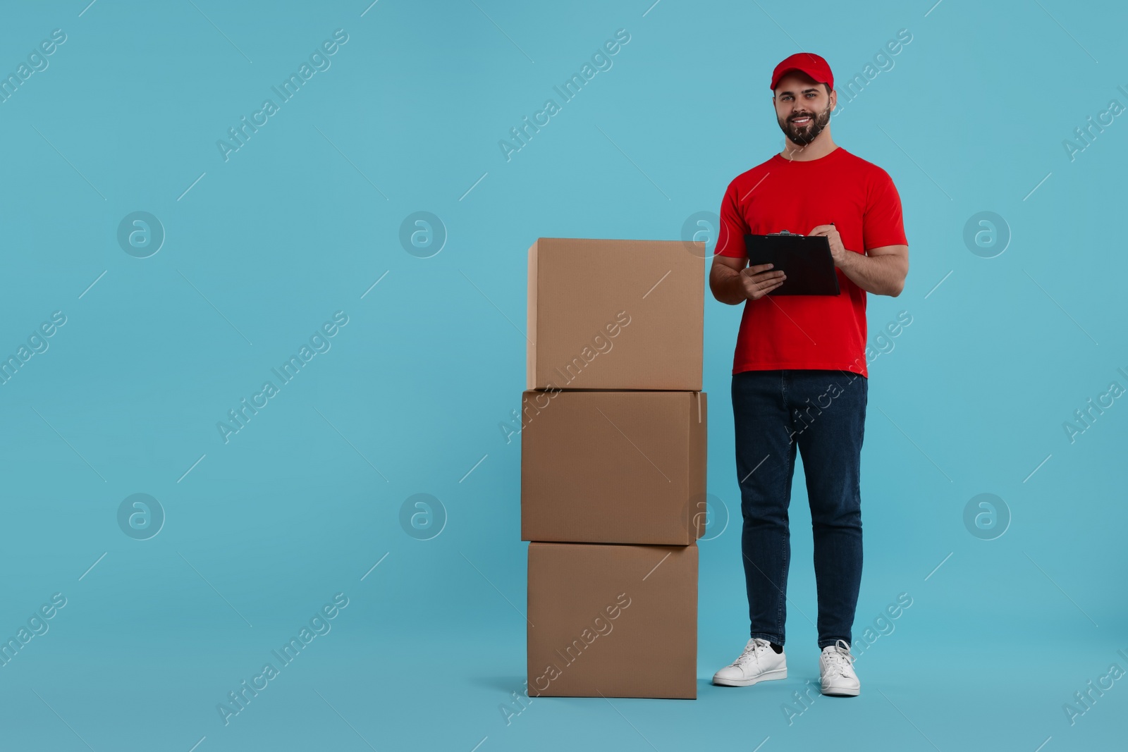 Photo of Happy young courier with clipboard and stack of parcels on light blue background, space for text