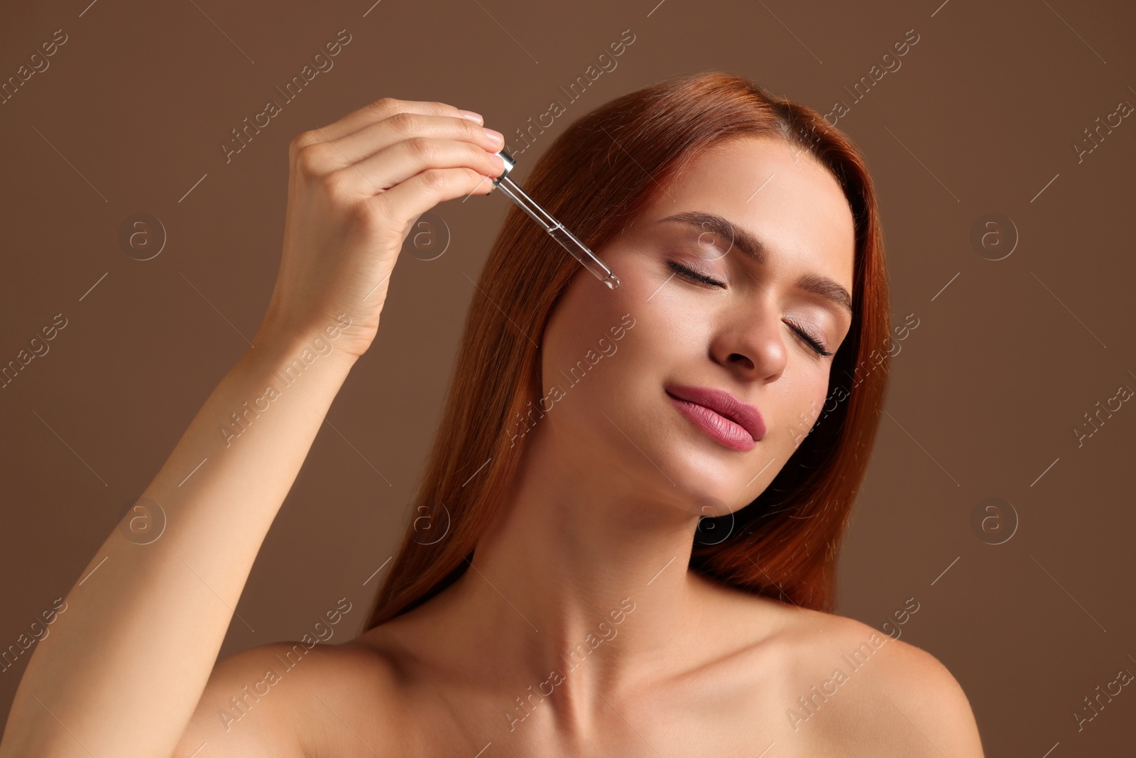 Photo of Beautiful young woman applying cosmetic serum onto her face on brown background