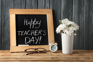 Photo of Chalkboard with inscription HAPPY TEACHER'S DAY and vase of flowers on wooden table against grey wall
