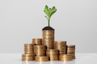Photo of Stacks of coins with green sprout on white table against light grey background. Investment concept