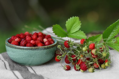 Bowl and tasty wild strawberries on cloth against blurred background