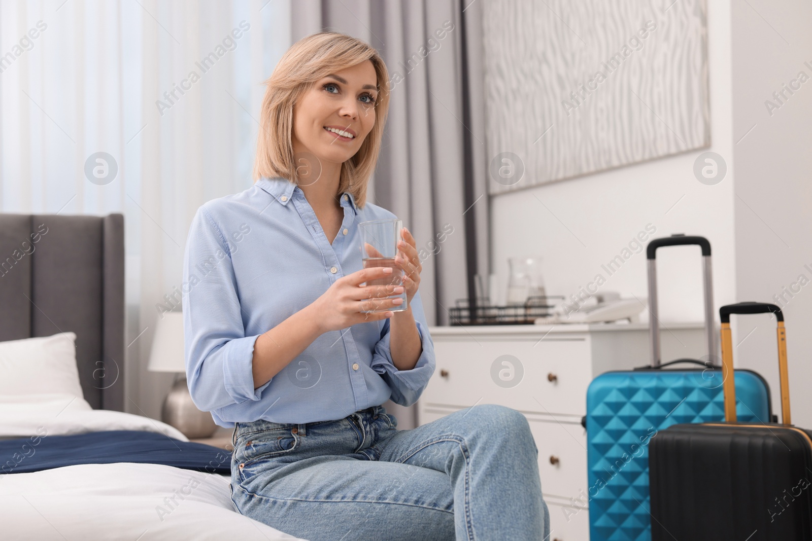 Photo of Smiling guest with glass of water on bed in stylish hotel room