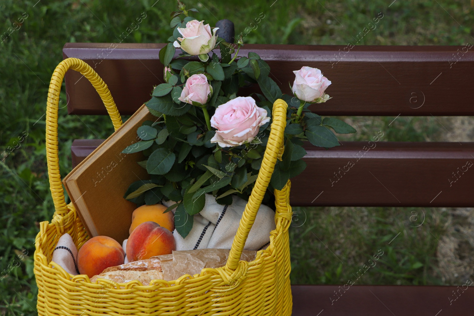 Photo of Yellow wicker bag with roses, book and peaches on wooden bench outdoors