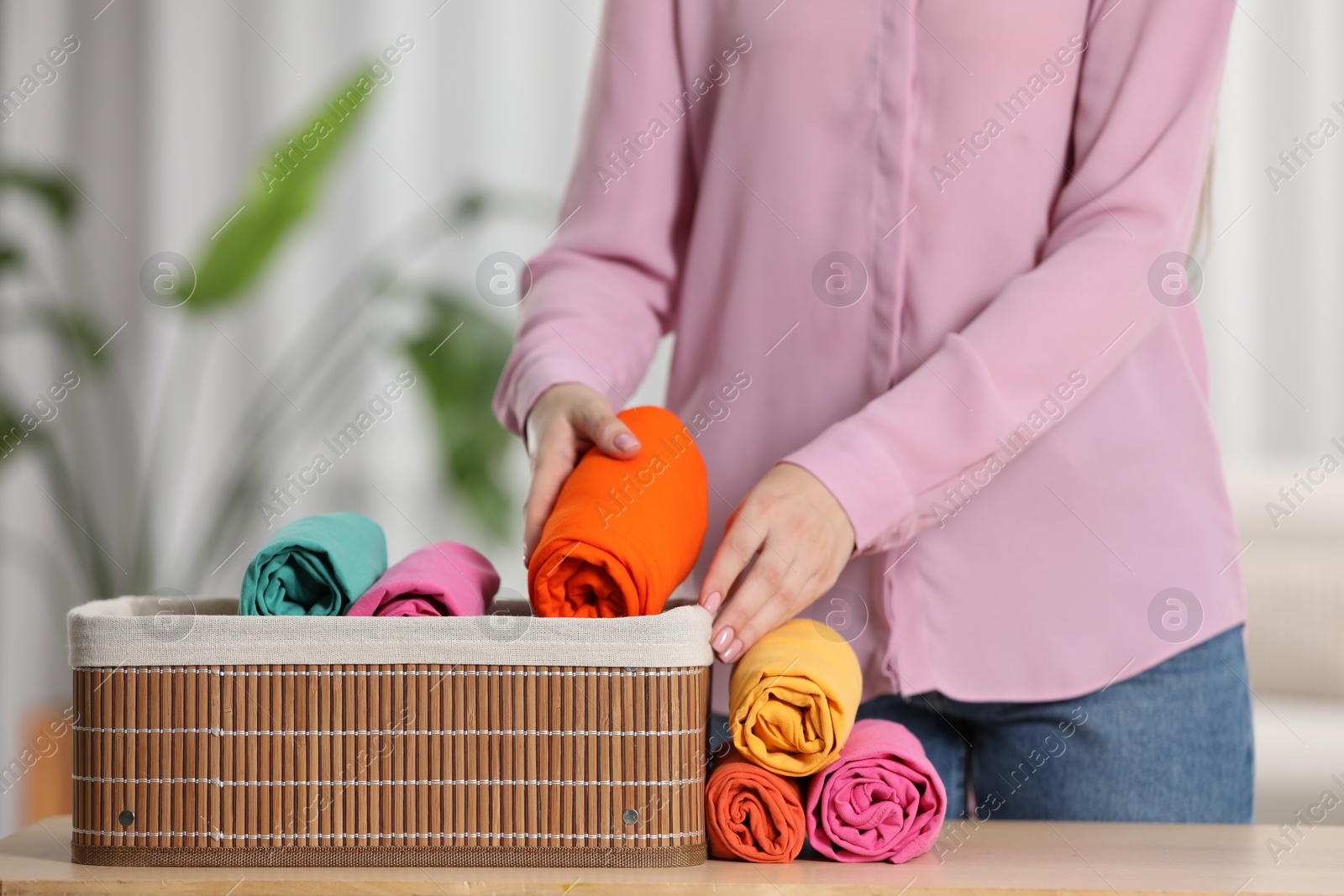 Photo of Woman putting rolled shirt into basket at table indoors, closeup. Organizing clothes