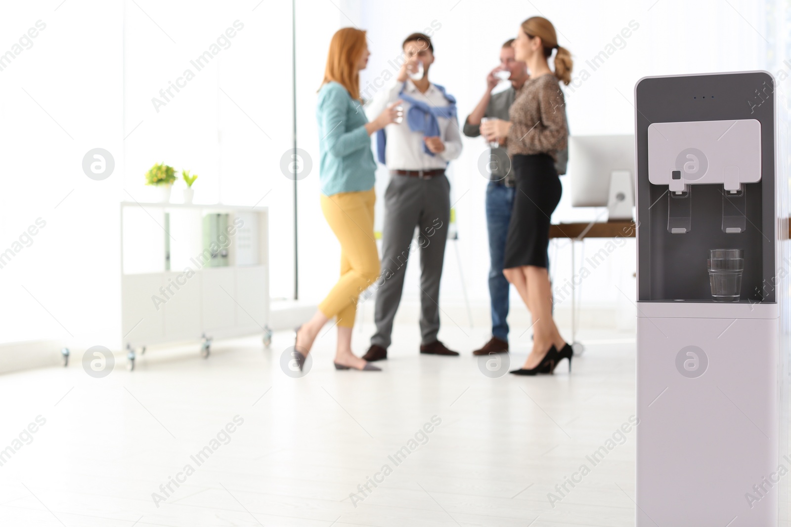 Photo of Modern water cooler with glass and blurred office employees on background. Space for text