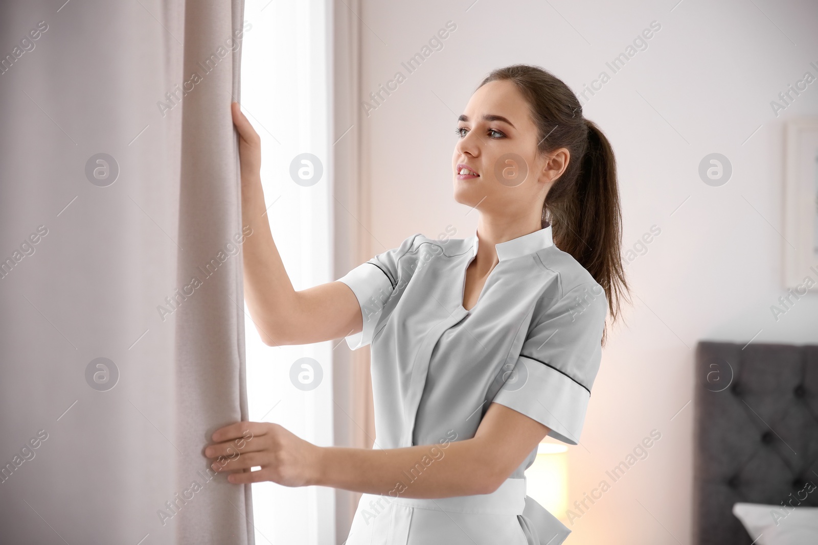 Photo of Young maid adjusting curtains in hotel room