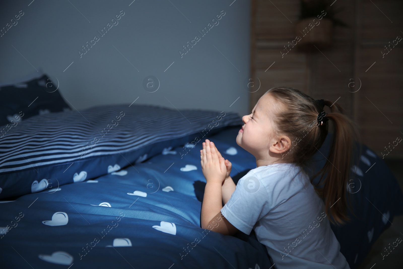 Photo of Little girl saying bedtime prayer near bed in room at night