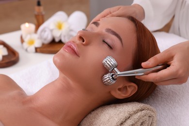 Photo of Young woman receiving facial massage with metal roller in beauty salon, closeup