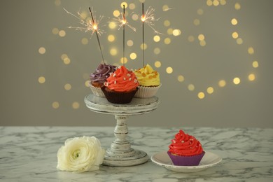 Photo of Different colorful cupcakes with sparklers on white marble table against blurred lights