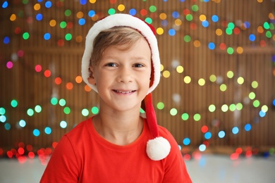 Photo of Cute little child in Santa hat on blurred lights background. Christmas celebration