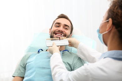 Dentist matching young man's teeth color with palette in office