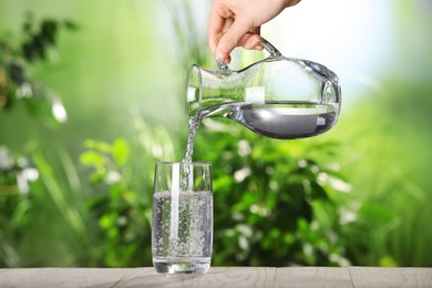 Photo of Woman pouring water from jug into glass on grey wooden table outdoors, closeup