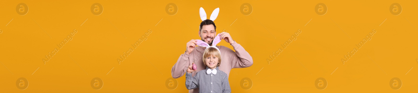 Image of Father and son in bunny ears headbands having fun on orange background, banner design. Easter celebration