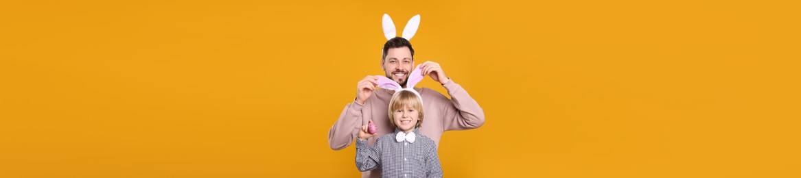 Image of Father and son in bunny ears headbands having fun on orange background, banner design. Easter celebration