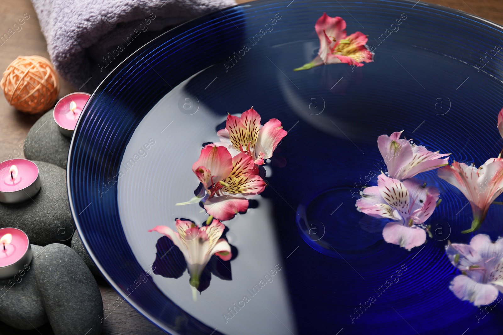 Photo of Bowl of water with flowers, candles, towel and stones on table, closeup. Spa treatment