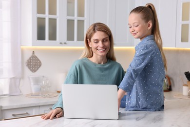 Happy woman and her daughter with laptop at white table indoors